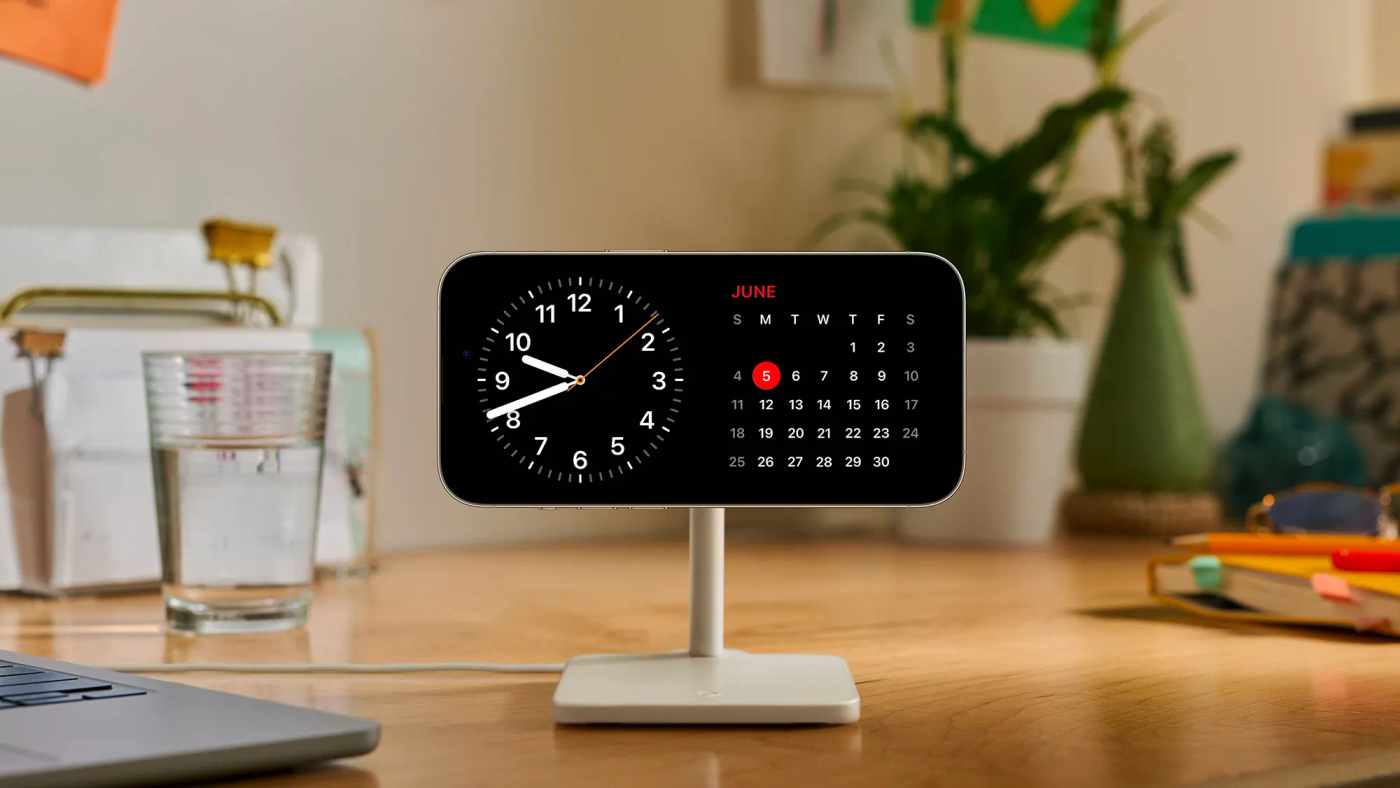 iPhone im StandBy-Modus auf einem Ständer: links eine analoge Uhr, rechts ein Kalender-Widget
